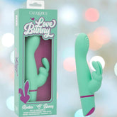 A turquoise silicone CalExotics Love Bunny Rockin’ “G” Bunny vibrator with a bunny-shaped clitoral stimulator is displayed beside its packaging on a soft, pastel background.